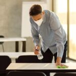 Mid adult teacher with face mask cleaning tables with disinfectant in the classroom.